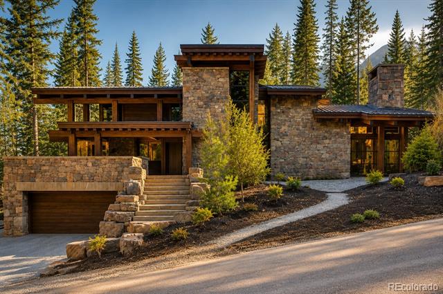 Modern stone home with an attached garage, landscaped entrance, and evergreen forest backdrop.