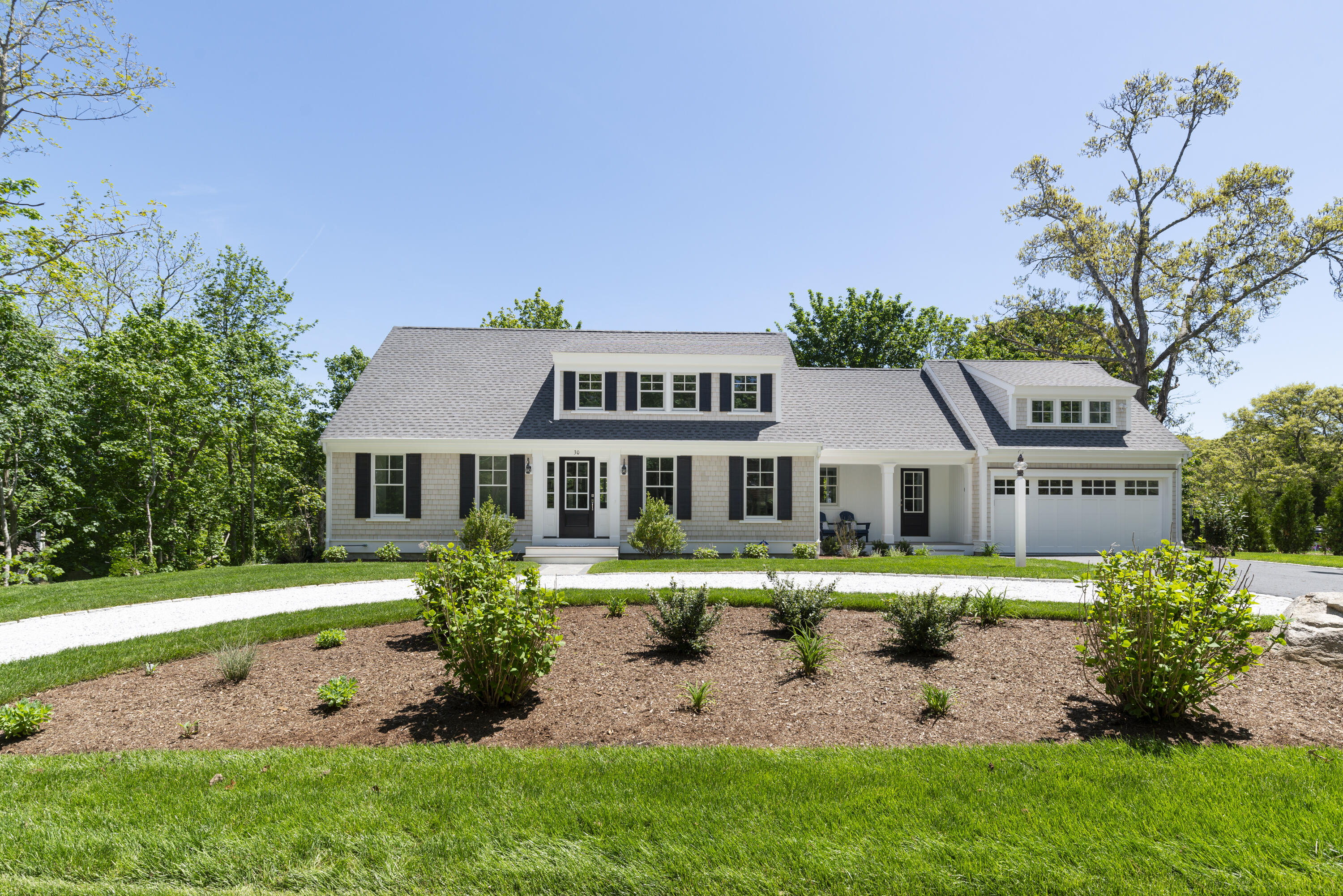 Light-gray shingle home with dormers, an attached garage, and a curved driveway through a landscaped lawn.