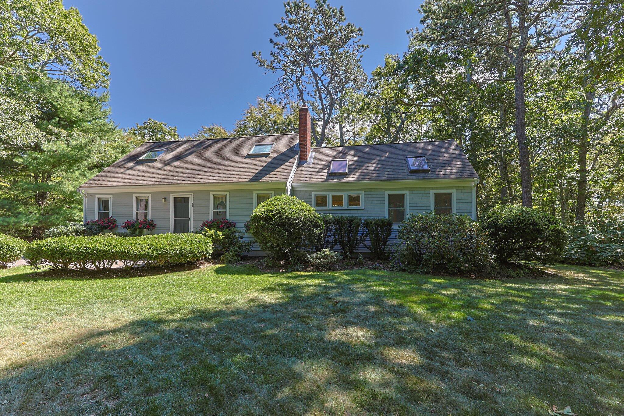 Gray house with dormer windows and brick chimney, surrounded by mature trees and a landscaped lawn.