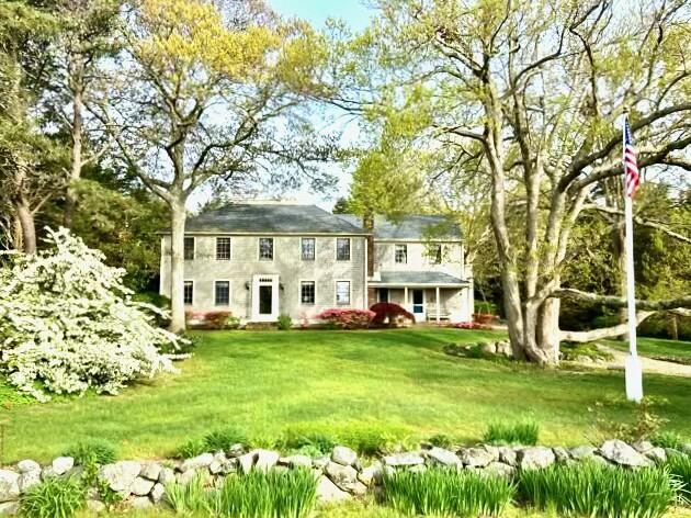 Two-story colonial-style home beyond a green lawn, framed by mature trees and a low stone wall.