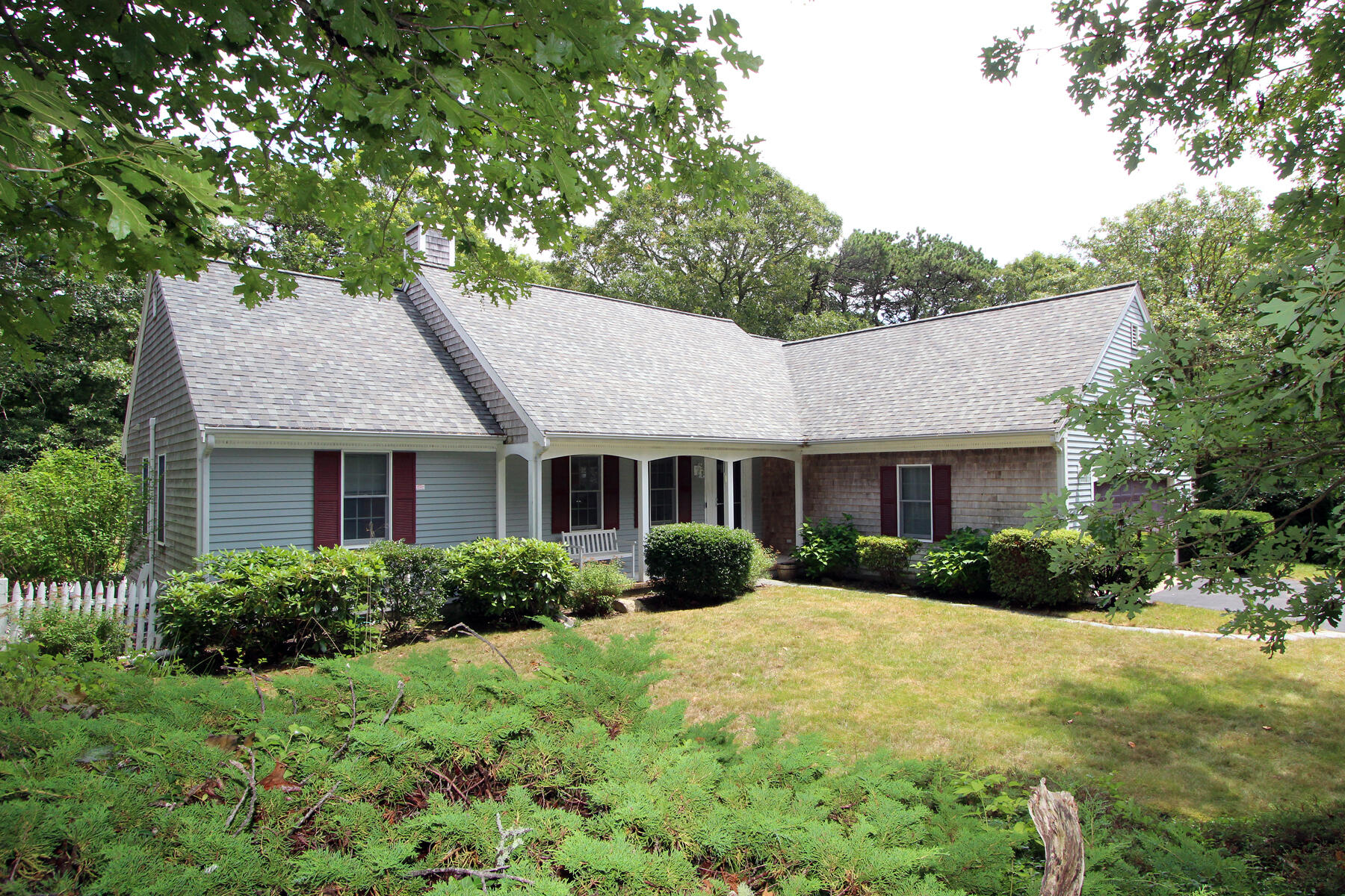 Gray Cape-style house with a covered porch, burgundy shutters, and a leafy landscaped yard.