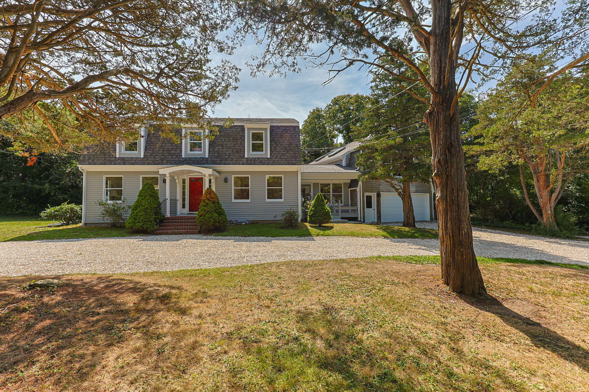 Gray-shingled two-story house with a red front door, attached garage, and mature trees framing the yard.