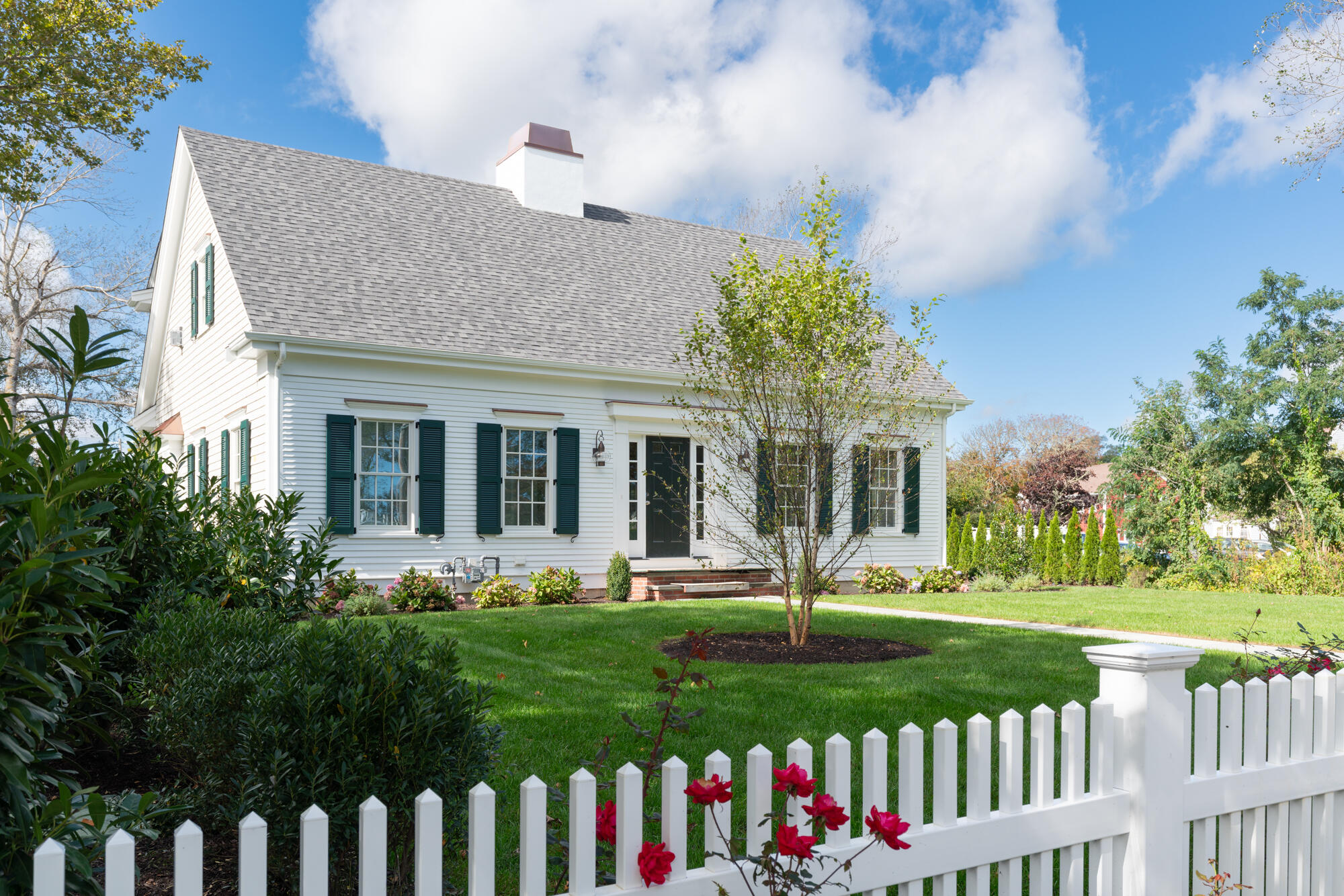 White clapboard house with dark green shutters, landscaped lawn, and white picket fence.