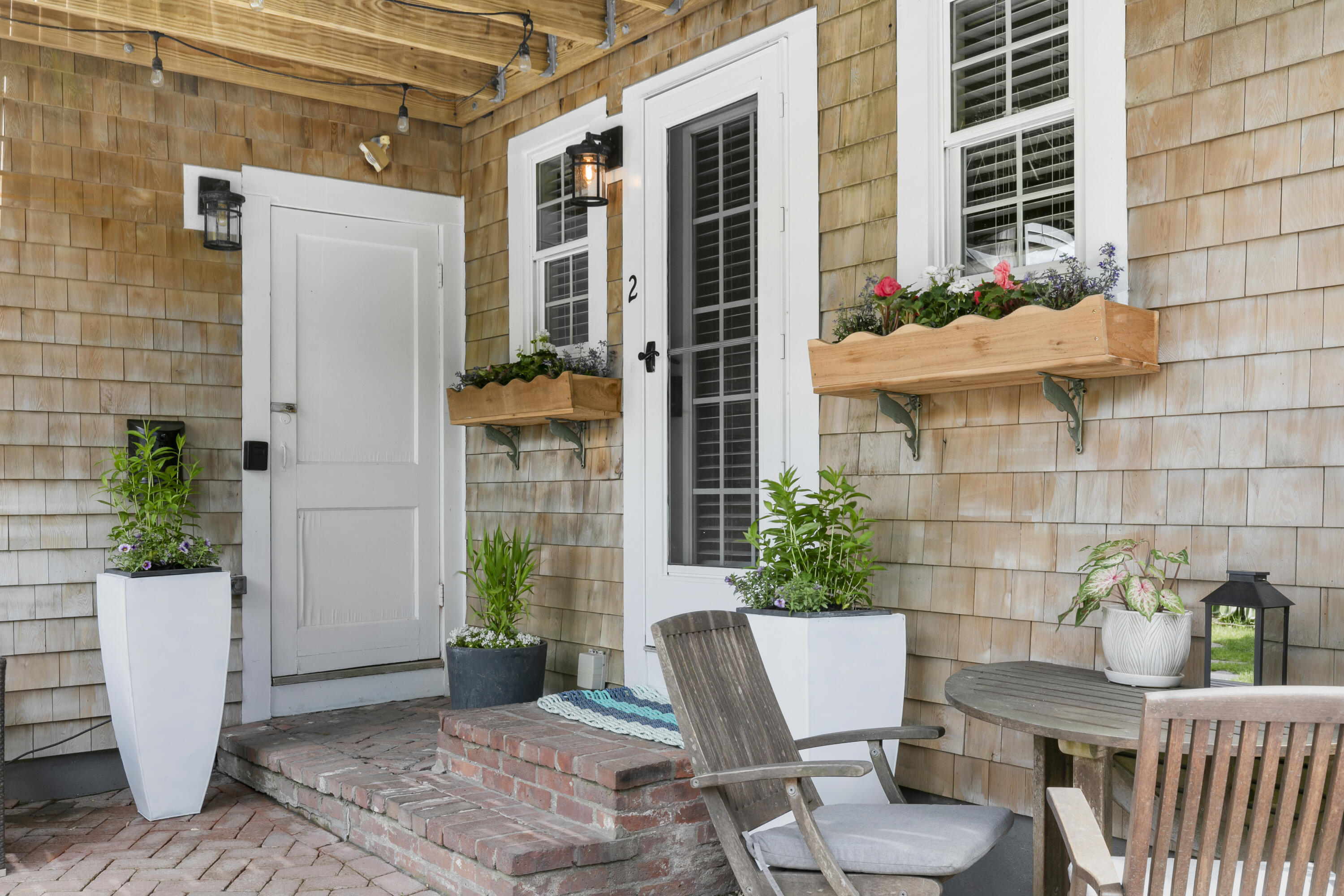 Covered cedar-shingle entry porch with brick steps, flower boxes, potted plants, and wooden seating.