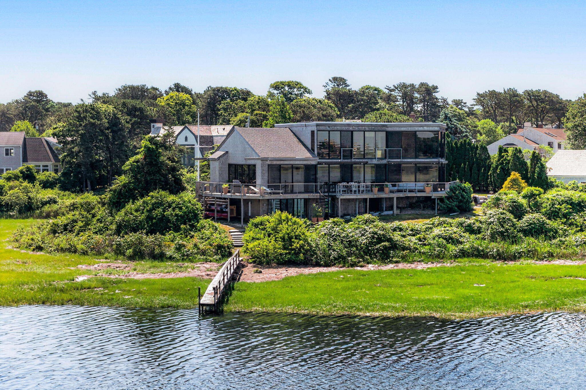 Modern glass-fronted waterfront home with a dock extending across the lawn to the water.