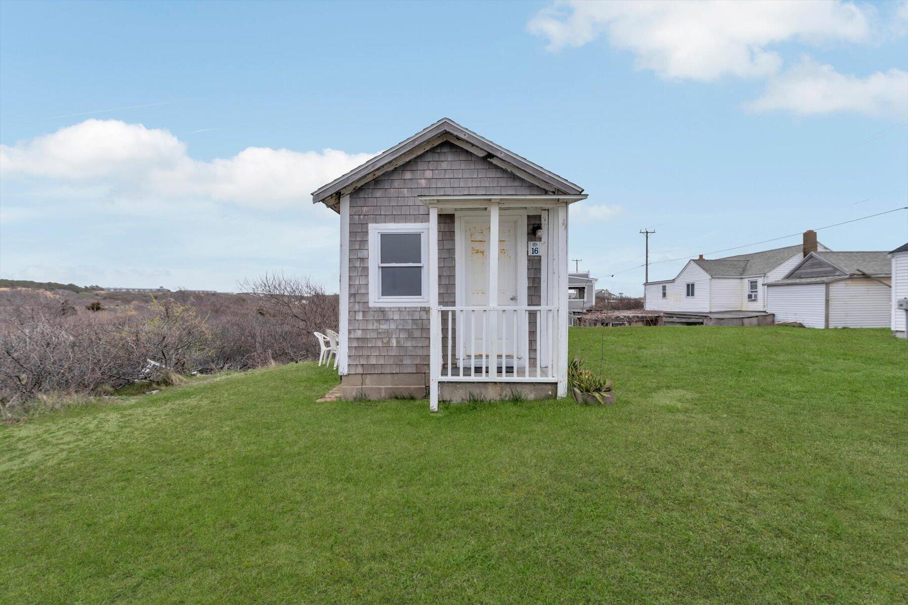 Small gray shingled building with a white porch and door marked 16 on a green lawn.