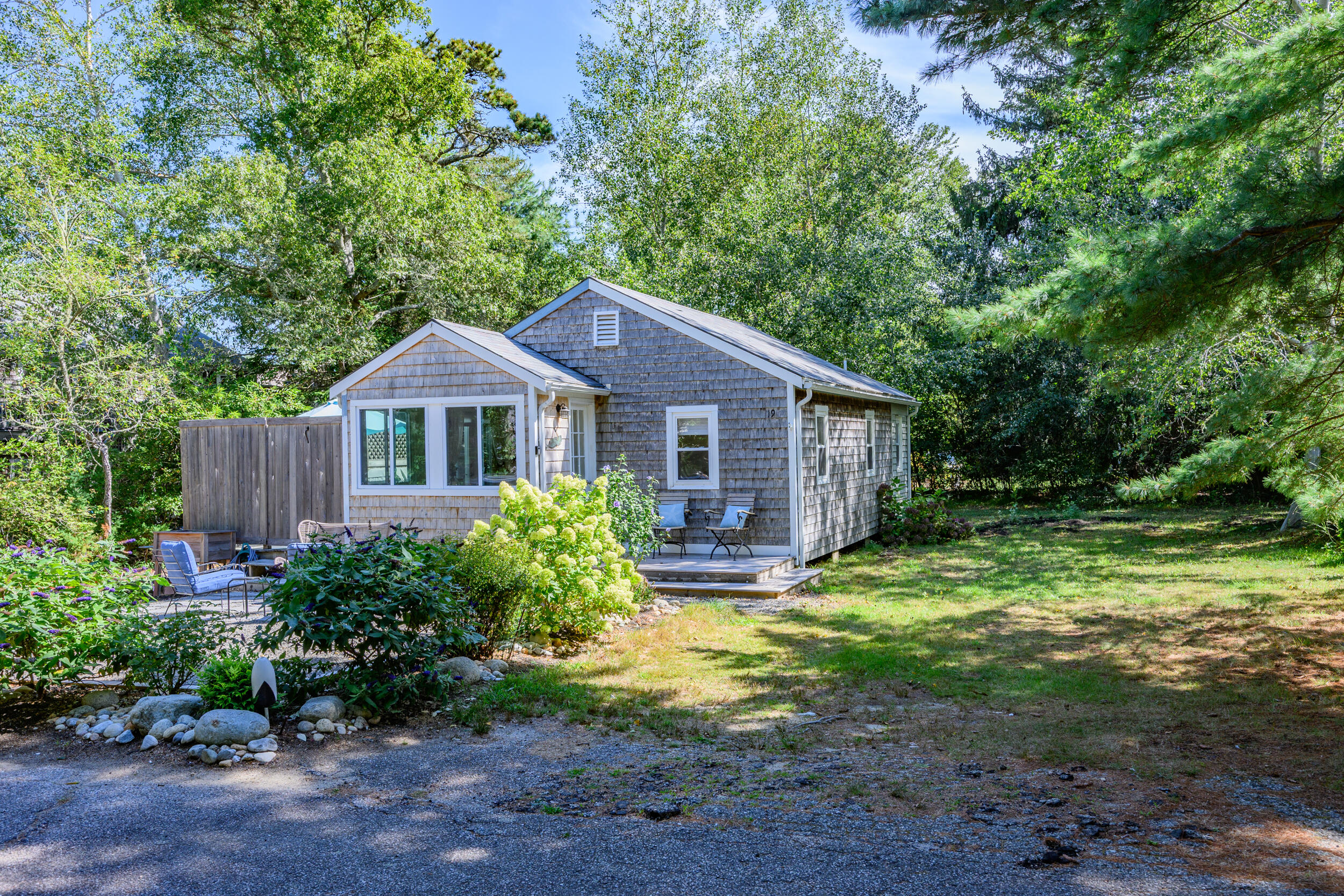 Gray shingle cottage nestled among trees, with a small deck, patio seating, and garden beds.