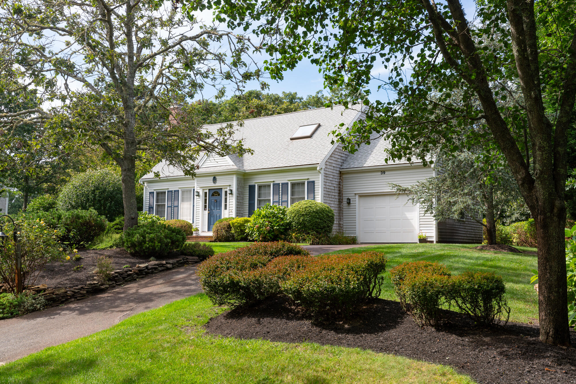 White house with blue shutters and door, attached garage, and landscaped front yard beneath mature trees.