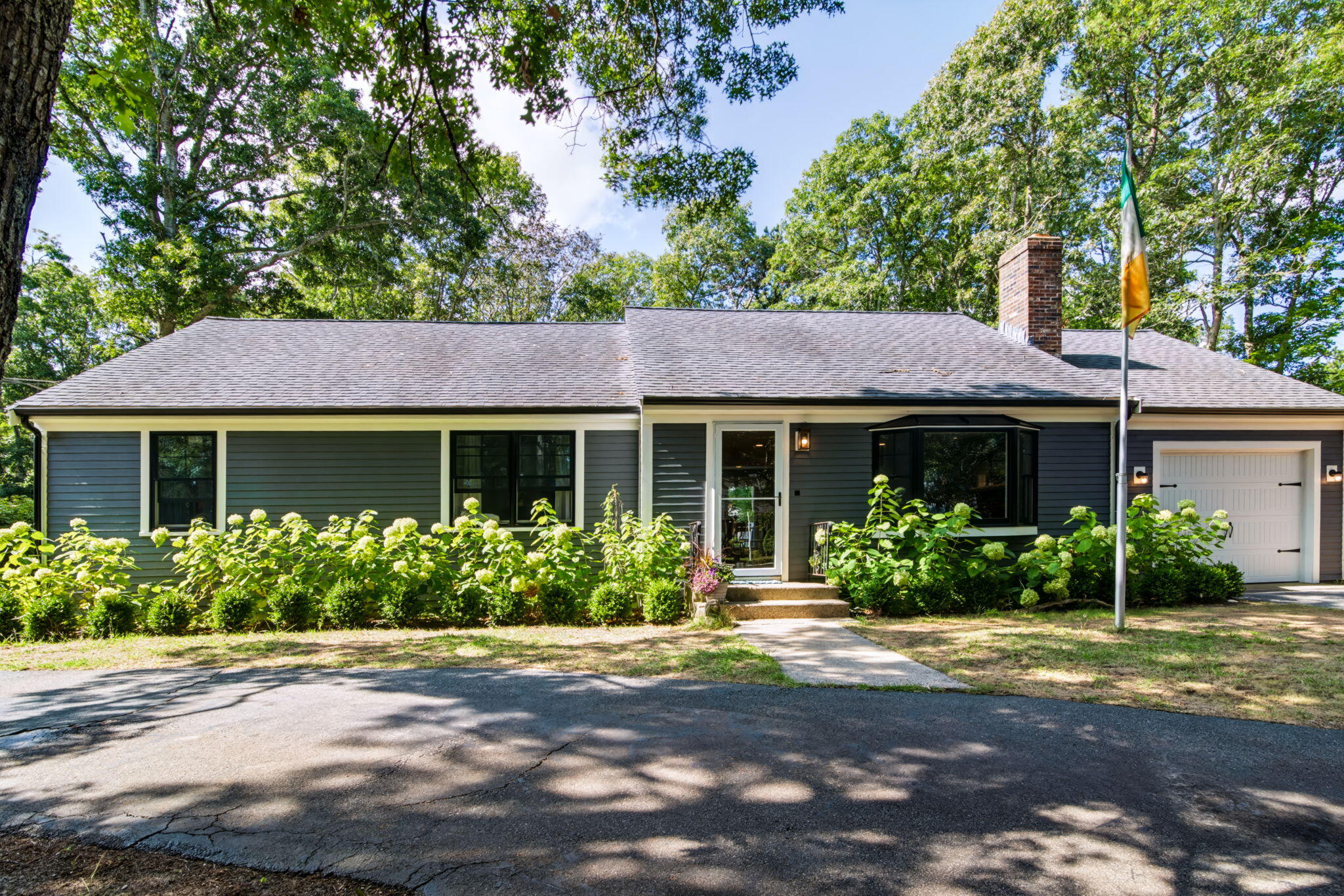 Gray single-story house with white trim, an attached garage, and flowering shrubs along the front.