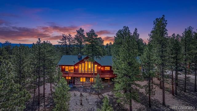 Multi-level home glowing among pine trees beneath a colorful dusk sky.