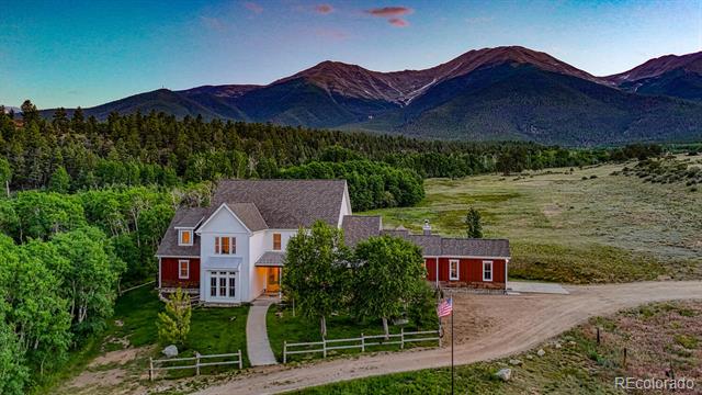 White and red farmhouse beside a gravel drive, with trees and mountains behind it.