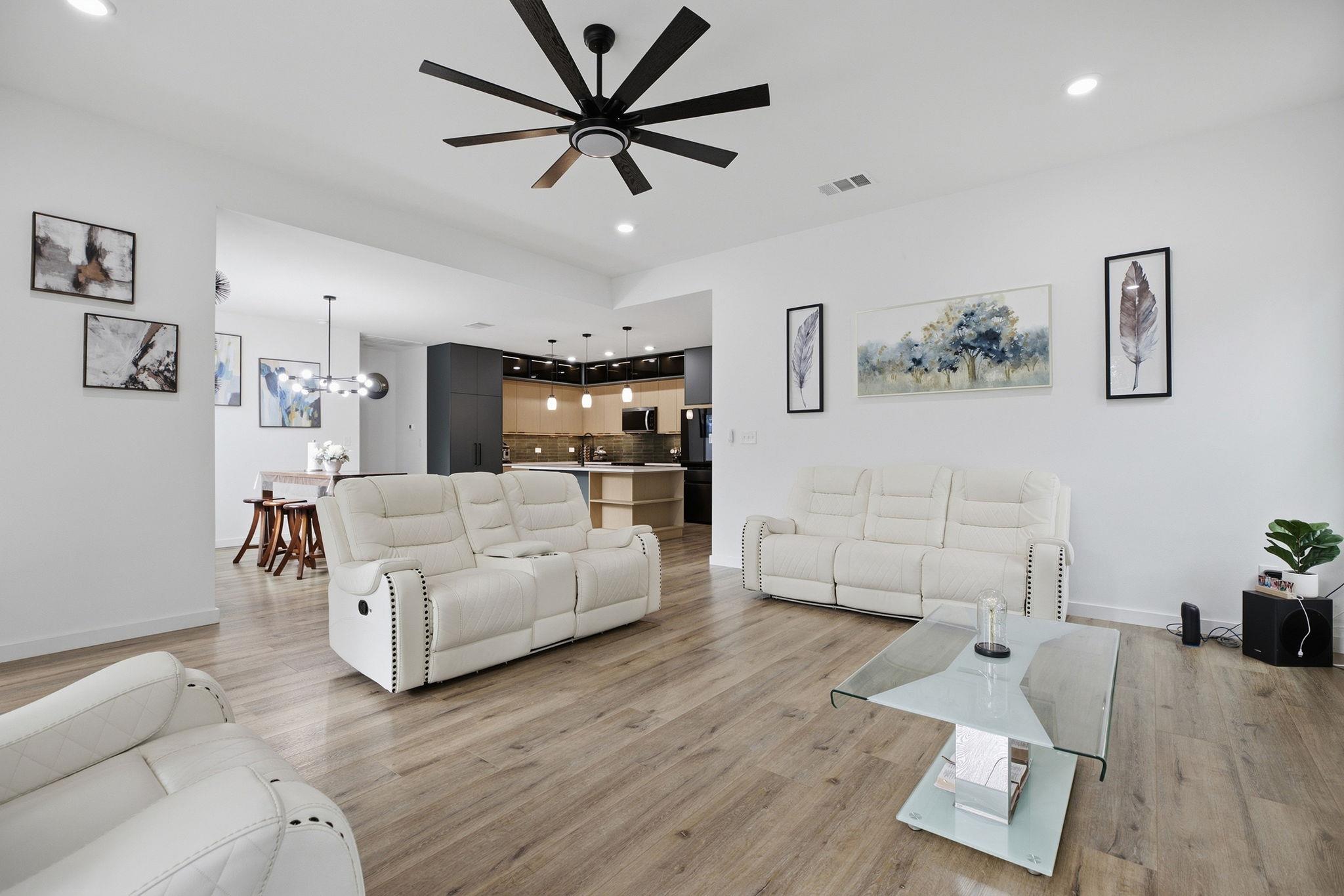 Open-plan living room with white reclining sofas, wood floors, and a modern kitchen beyond.