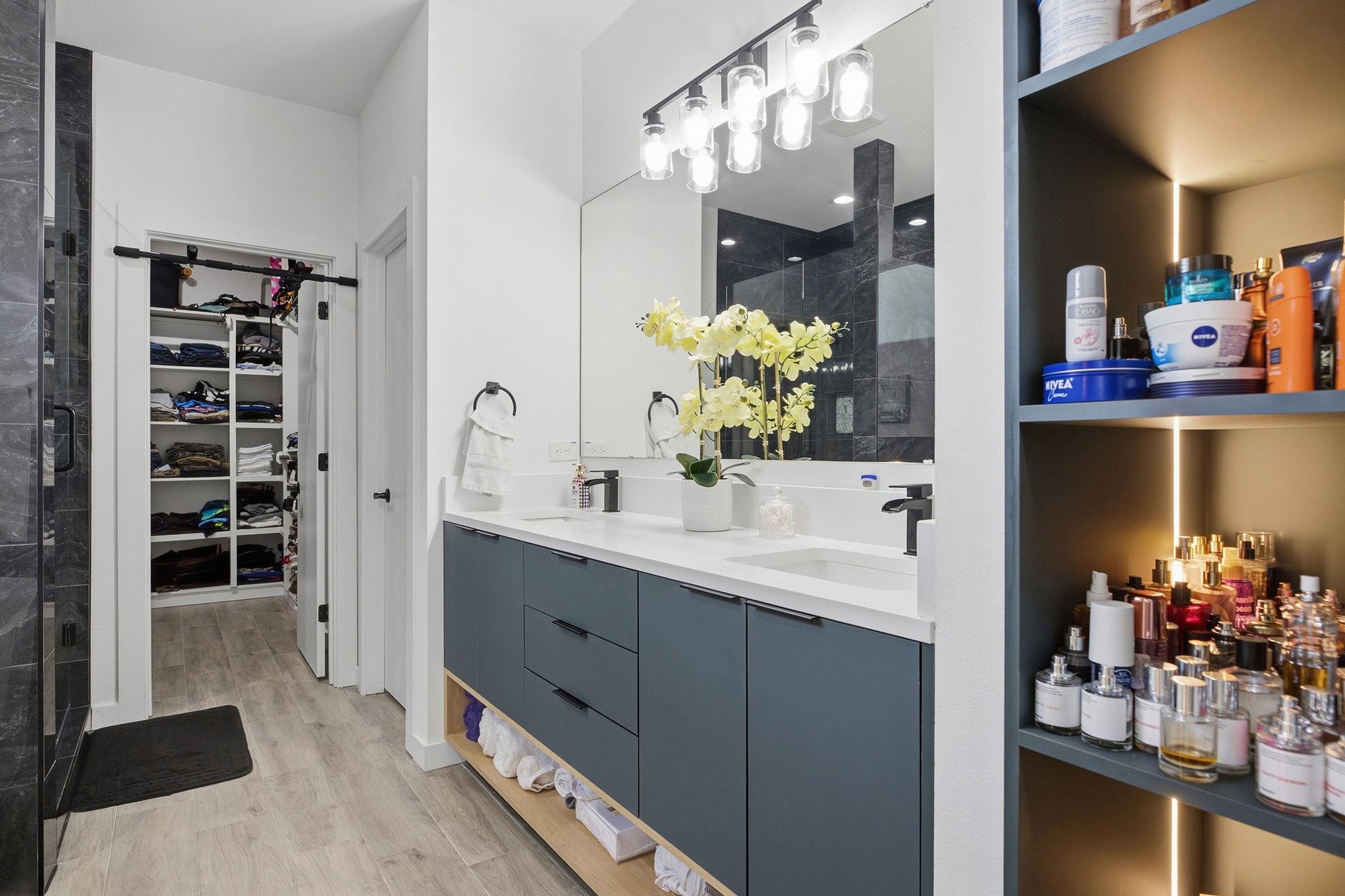 Modern bathroom with a double vanity, dark tiled shower, open linen closet, and illuminated toiletry shelves.