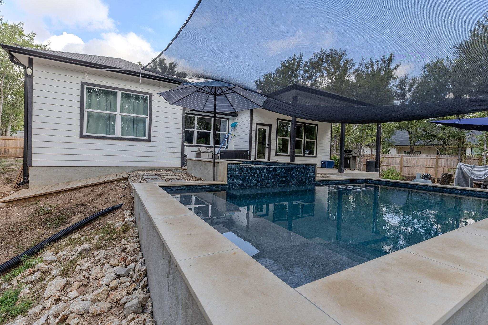 Backyard pool beside a white-sided home, shaded by a large sail and patio umbrella.