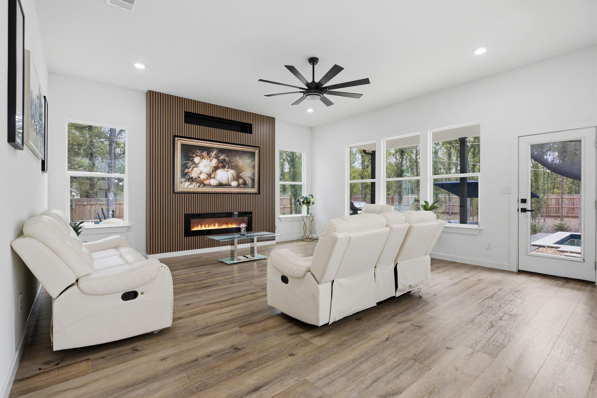 Living room with white reclining sofas, a slatted fireplace wall, and large windows overlooking trees.