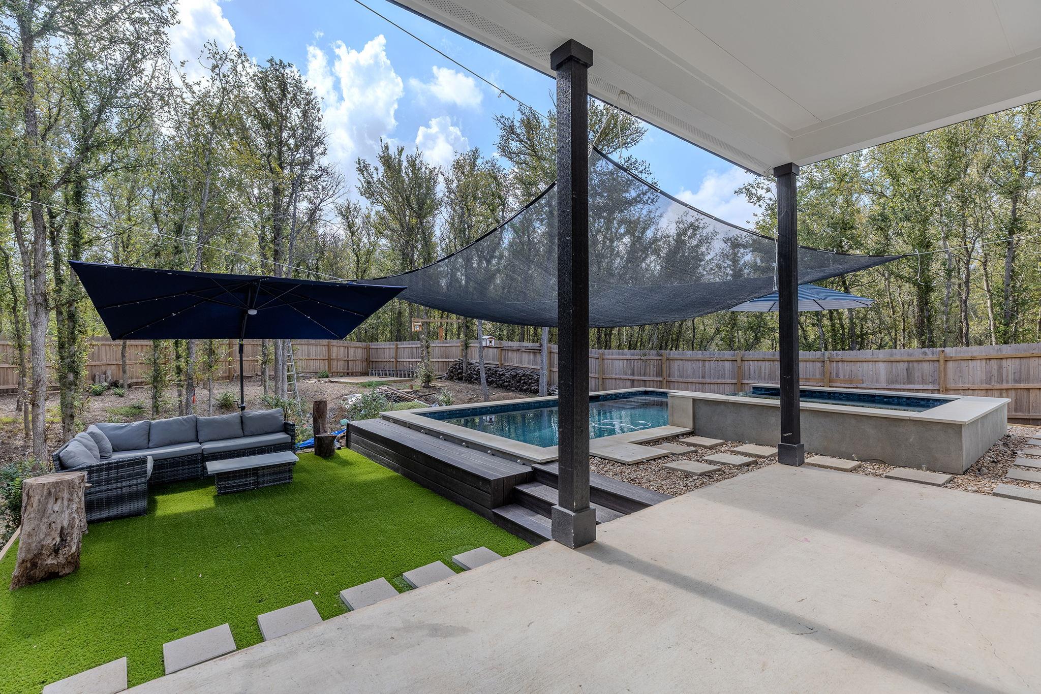 Backyard pool beside a covered patio, artificial turf, outdoor seating, and overhead shade sails.