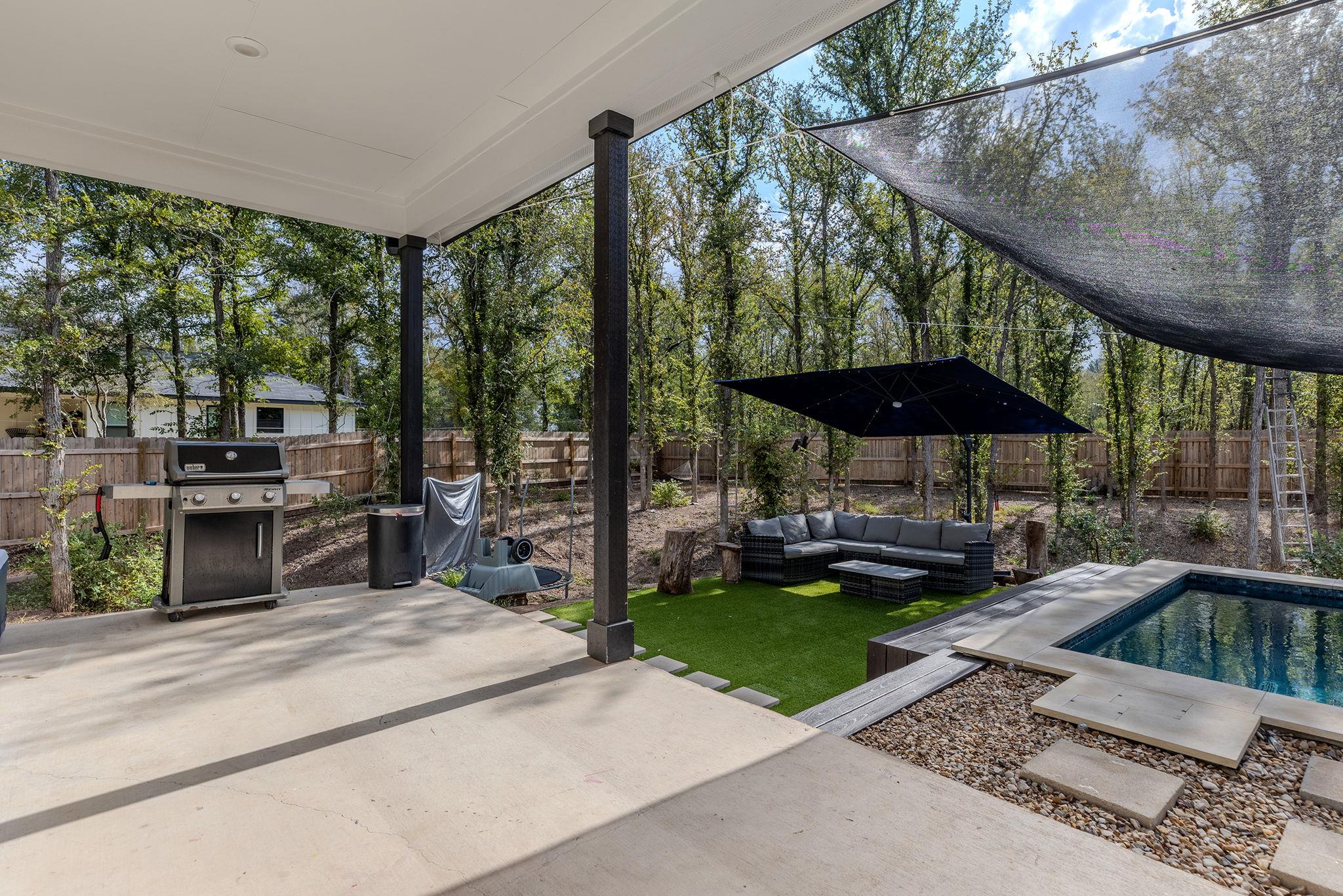 Covered patio beside a pool, with a barbecue grill and shaded outdoor seating in a fenced, wooded yard.