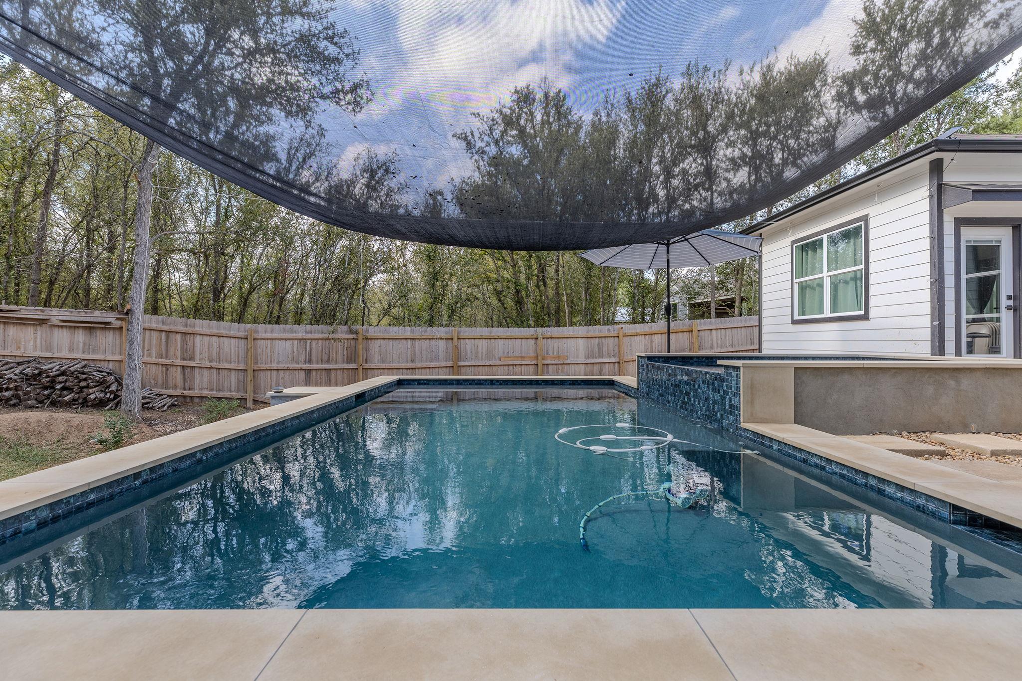 Backyard swimming pool beneath a shade sail, beside a white-sided house and fenced yard.
