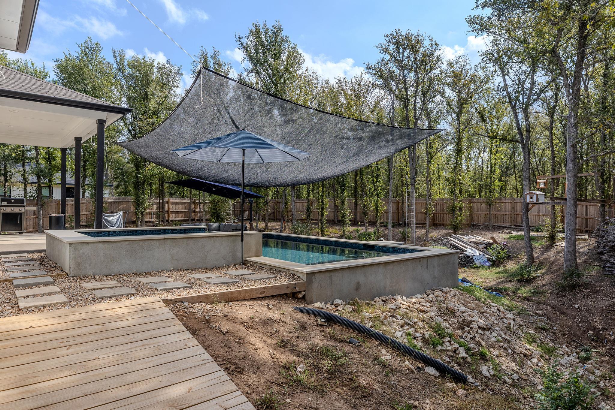 Backyard pool beneath a large shade sail, beside a wooden deck and wooded fenced yard.