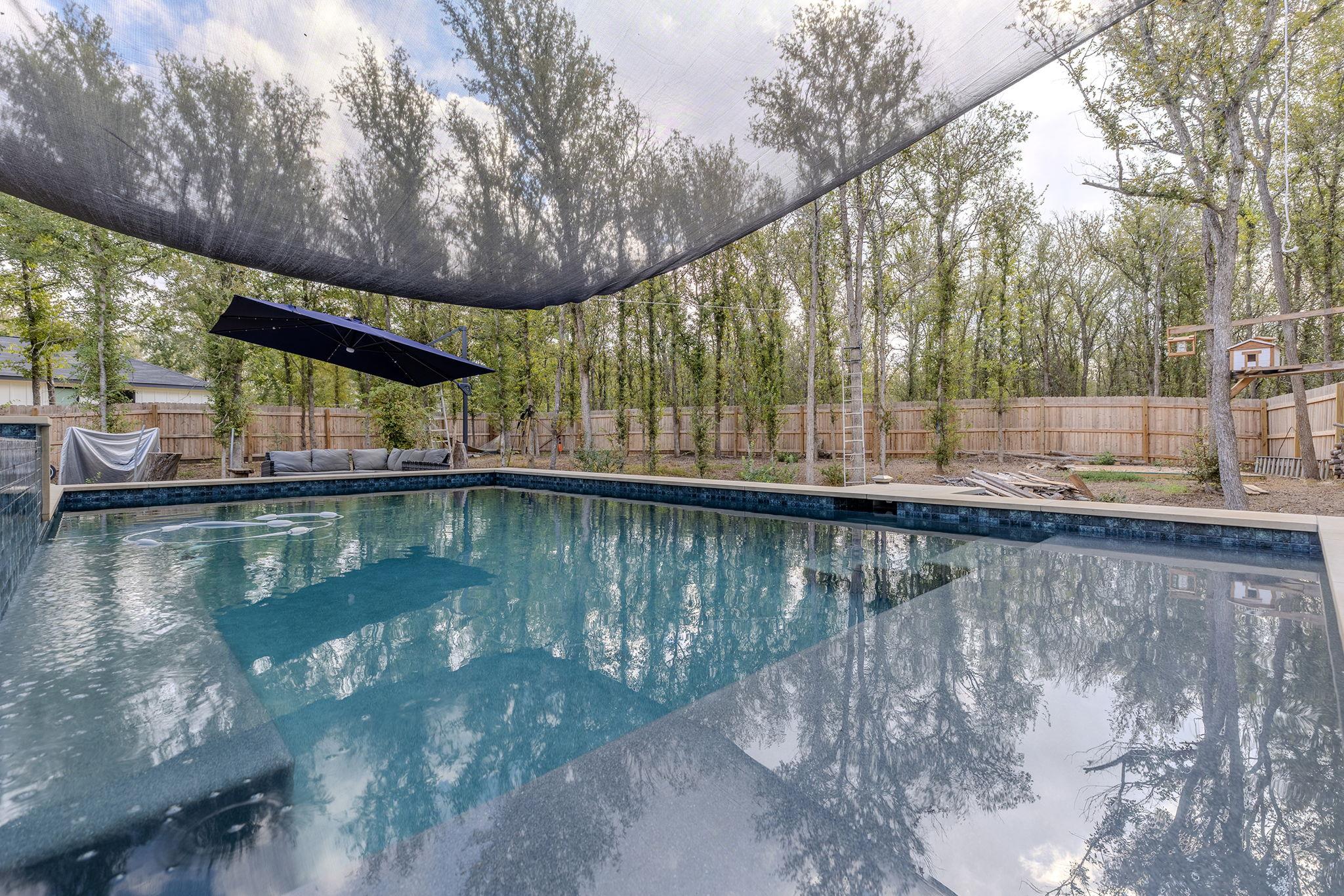 Backyard swimming pool beneath a black shade canopy, beside lounge seating and a wooded fenced yard.