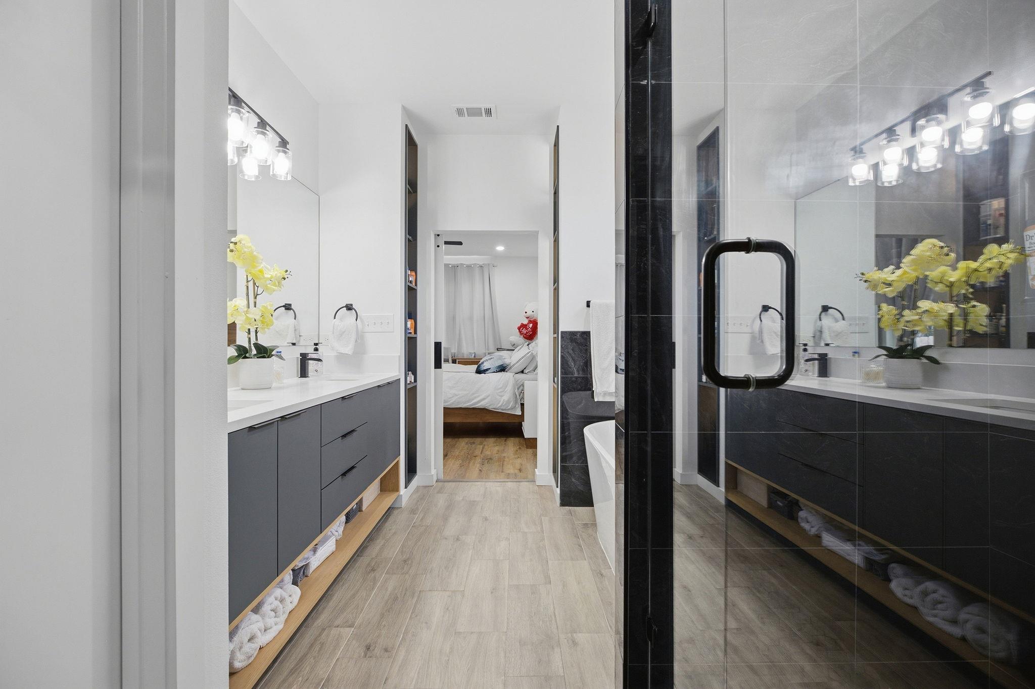Modern bathroom with a long double vanity and glass shower, viewed toward an adjoining bedroom.