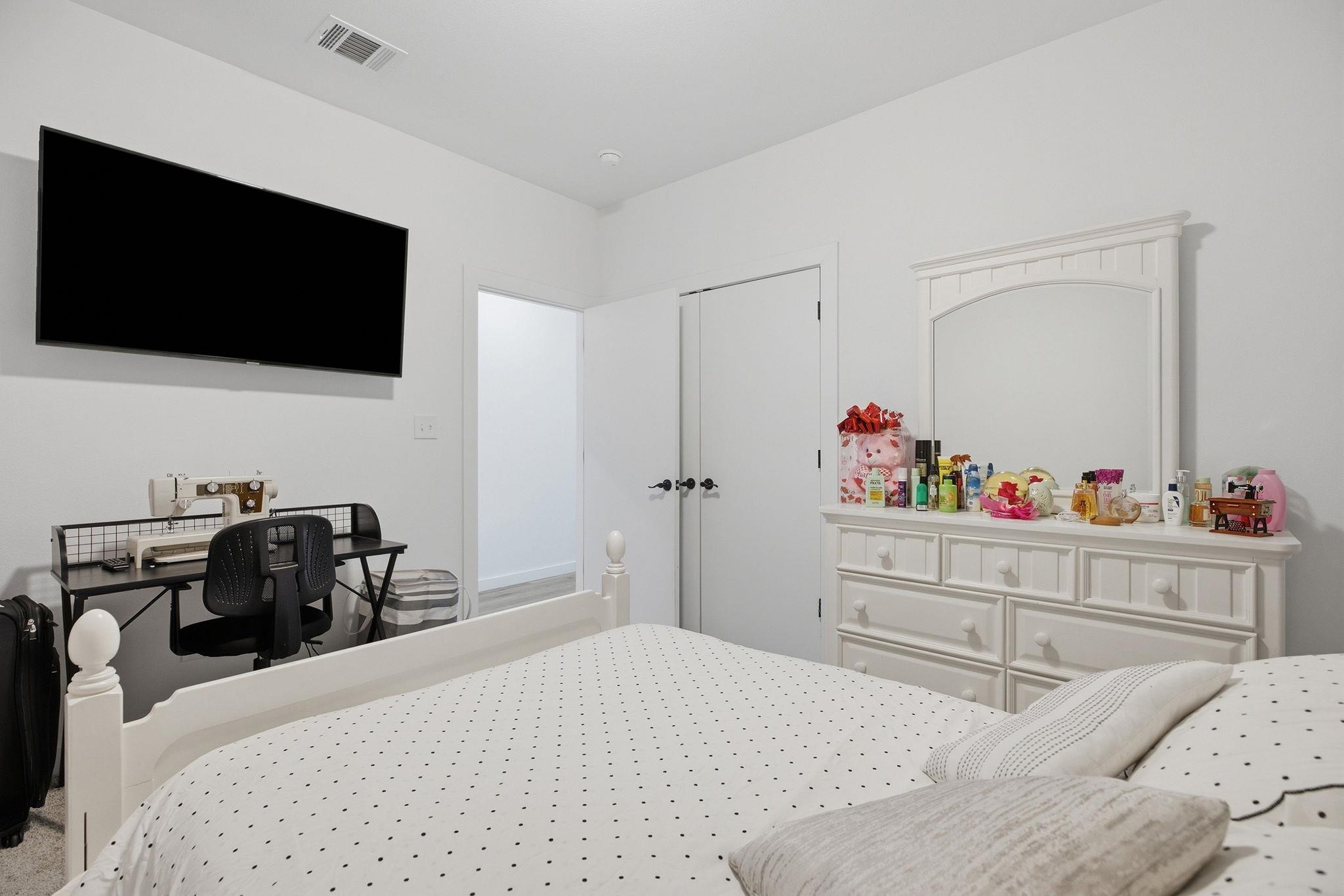Bedroom with dotted white bedding, a wall-mounted TV, sewing desk, and white dresser.