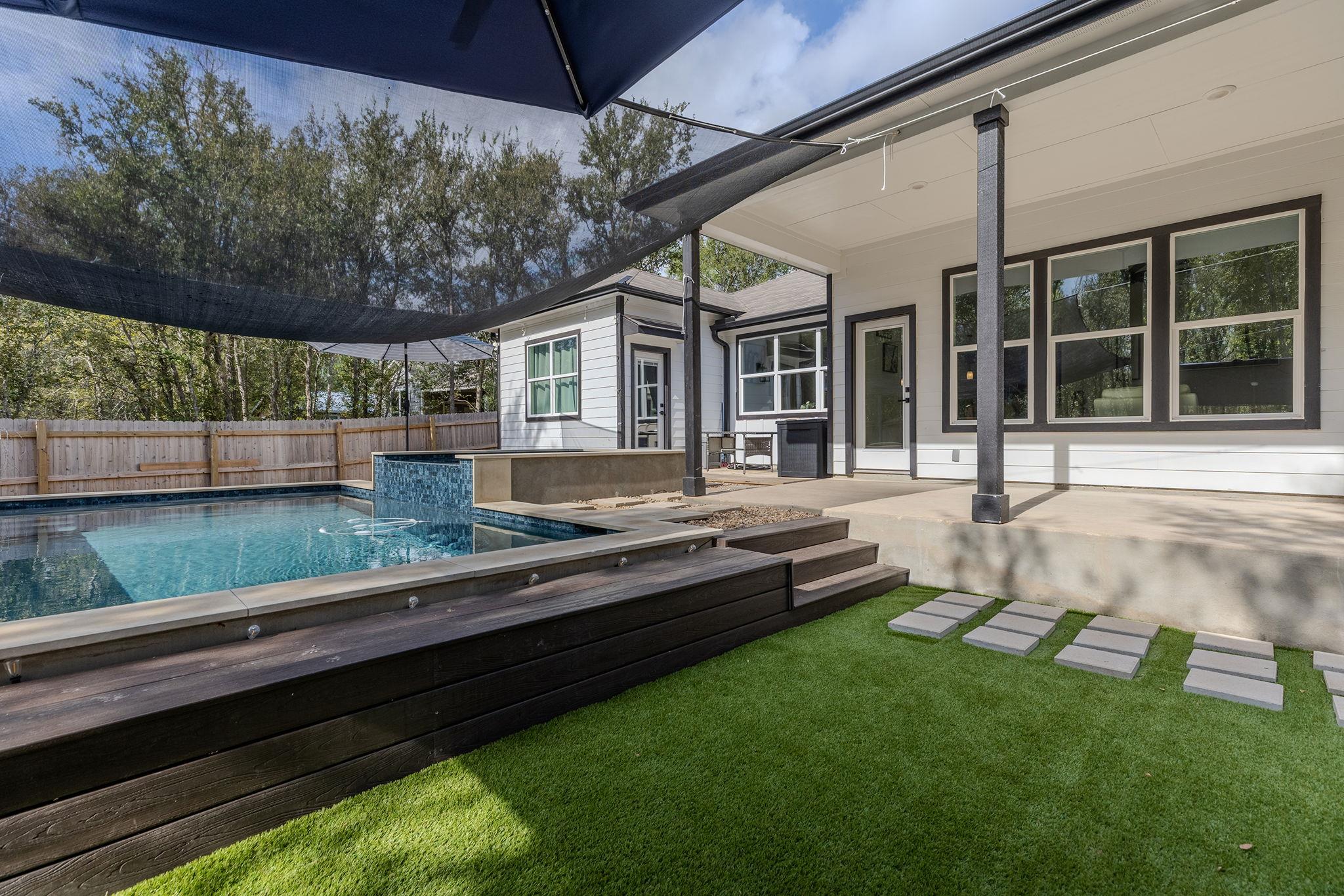 Backyard pool beside a covered patio, with dark wood decking, shade sails, and artificial turf.