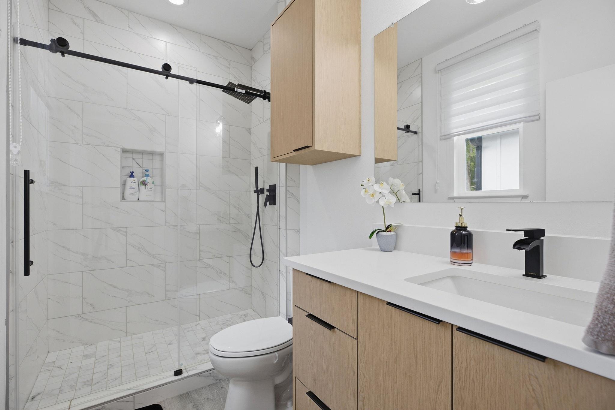 Modern bathroom with a marble-look tiled shower, glass enclosure, and light wood vanity.