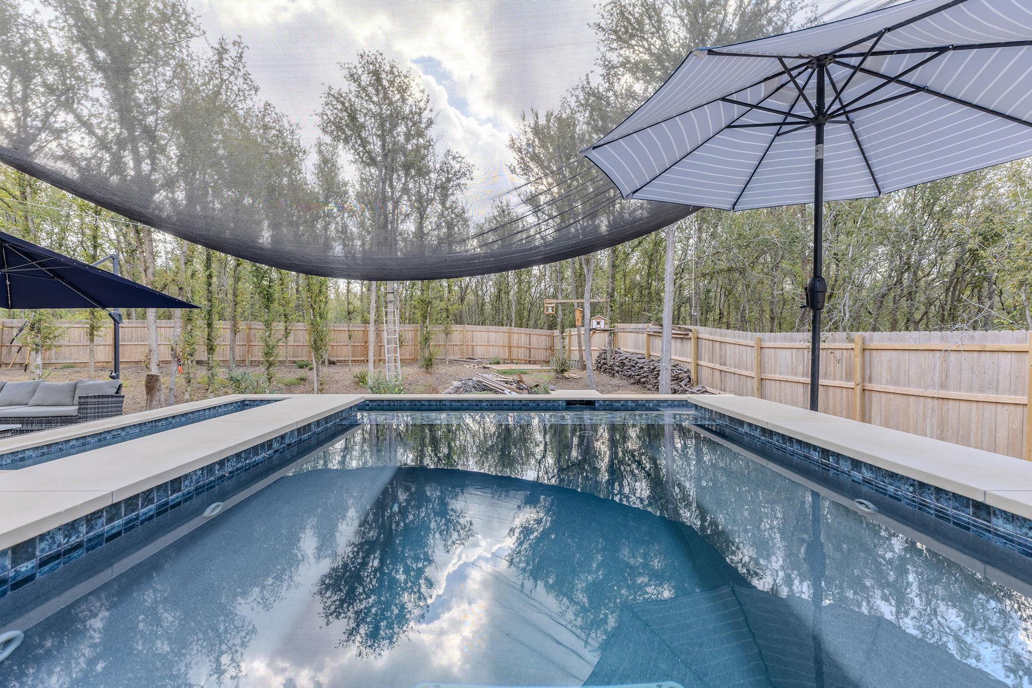 Backyard swimming pool beneath a large umbrella and shade canopy, enclosed by a wooden fence and trees.