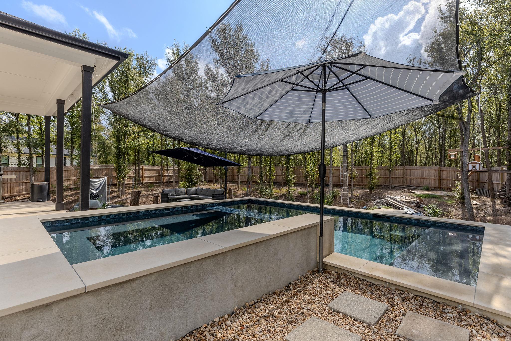 Backyard pool shaded by sails and umbrellas beside a fenced, wooded yard.
