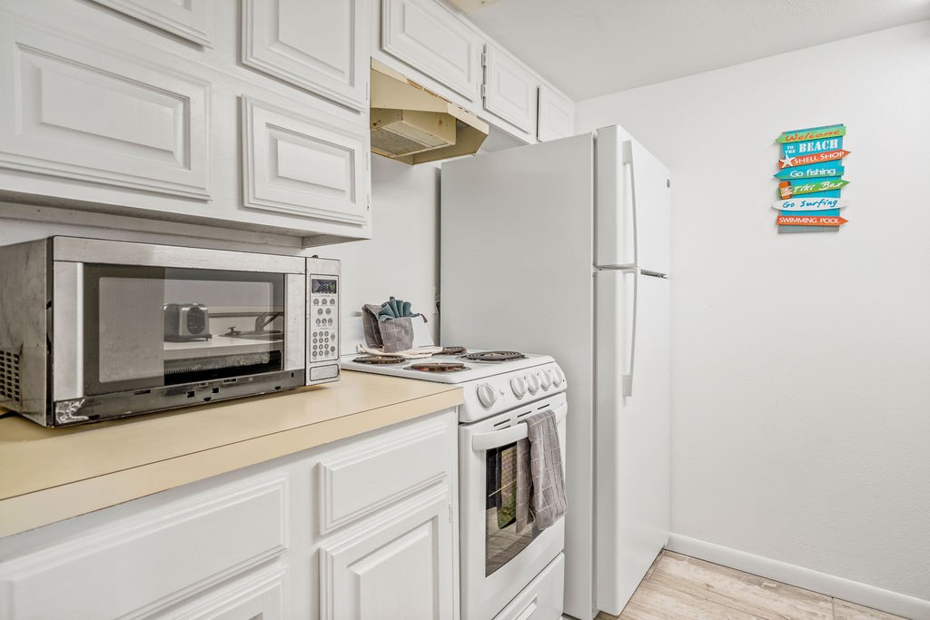 Compact white kitchen with a microwave, electric range, and refrigerator beside a beach-themed wall sign.