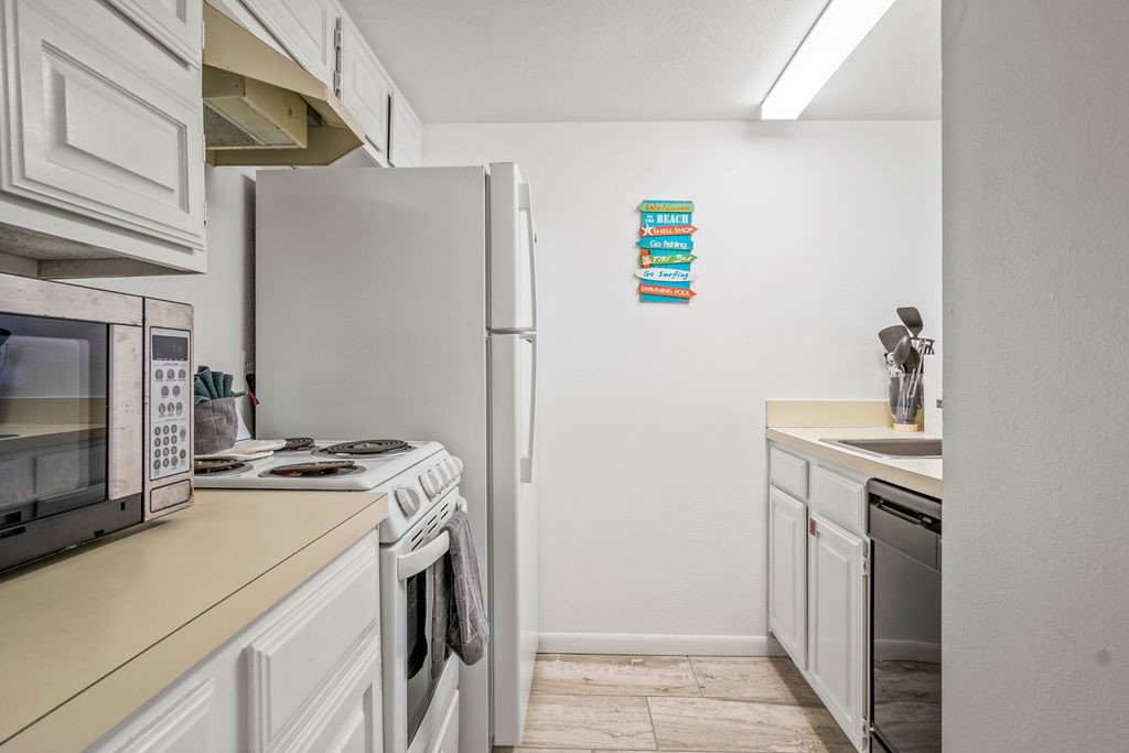 Compact kitchen with white cabinets, refrigerator, electric stove, and a colorful beach-themed sign.