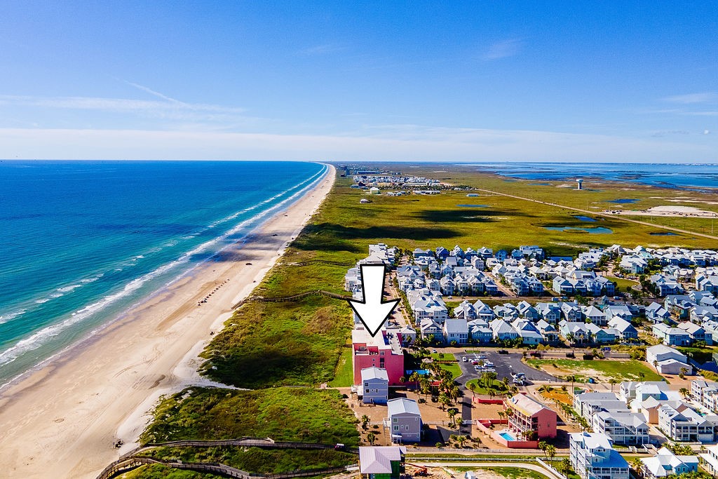Aerial view of a red-roofed beachfront building marked by an arrow, with ocean, sandy shore, and coastal homes.