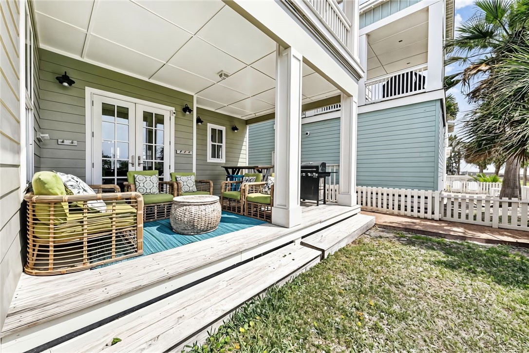 Covered patio with wicker seating, blue rug, barbecue grill, and white deck steps beside a grassy yard.