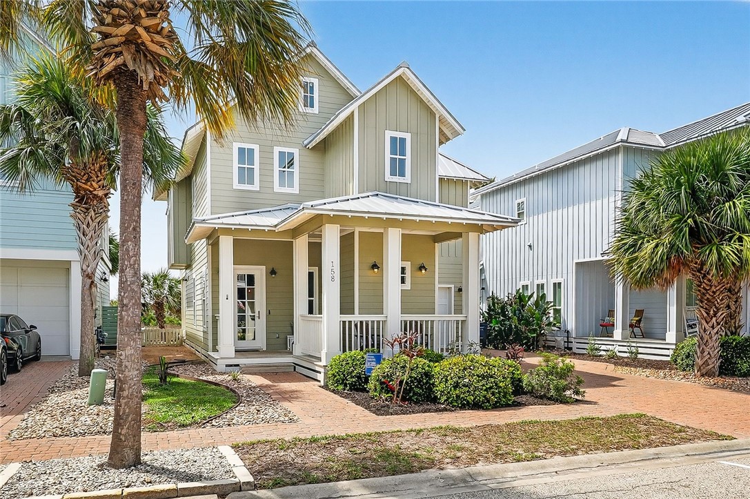 Two-story sage-green house with a covered porch, metal roof, and palm trees.