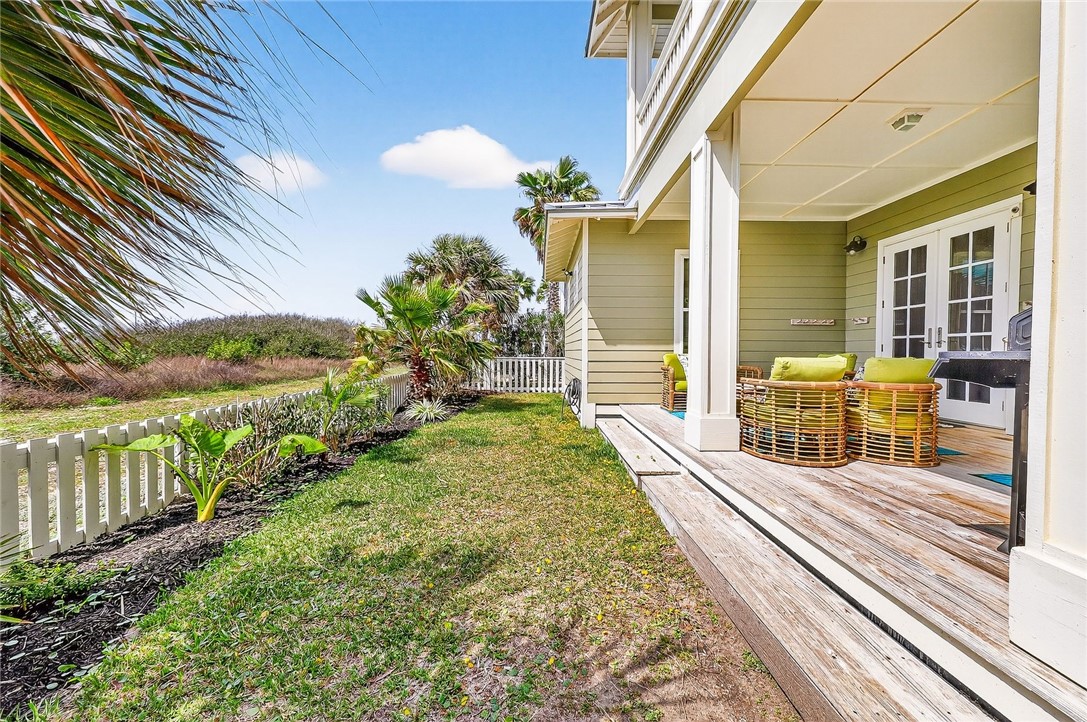 Home’s covered deck with wicker seating beside a grassy, palm-lined yard and white picket fence.