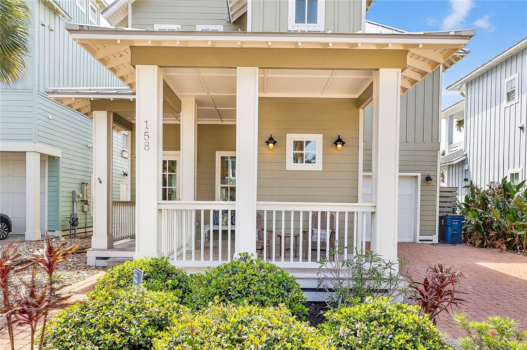 Coastal-style home with a broad covered porch, white railings, landscaped shrubs, and a brick-paved driveway.