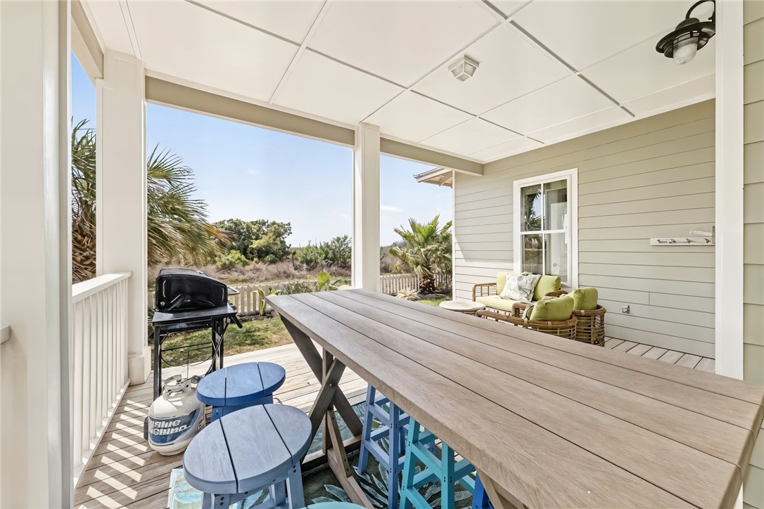 Covered patio with a long wooden dining table, grill, and cushioned seating overlooking palm trees.