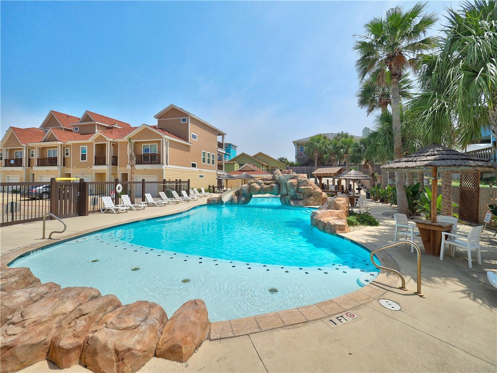 Condominium pool with a rock waterfall, shallow entry, palm trees, and shaded seating.