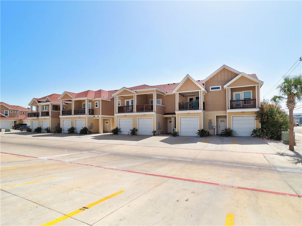 Row of tan condominiums with red roofs, balconies, and garage doors beside a marked concrete parking area.