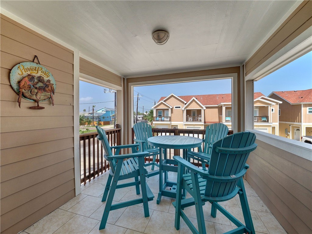 Covered tiled balcony with teal Adirondack chairs and a round table overlooking neighboring homes.