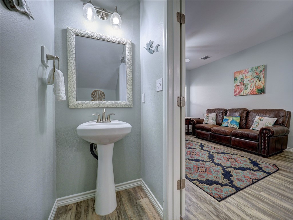 Bathroom pedestal sink and framed mirror beside an open doorway to a living room with a leather sofa.