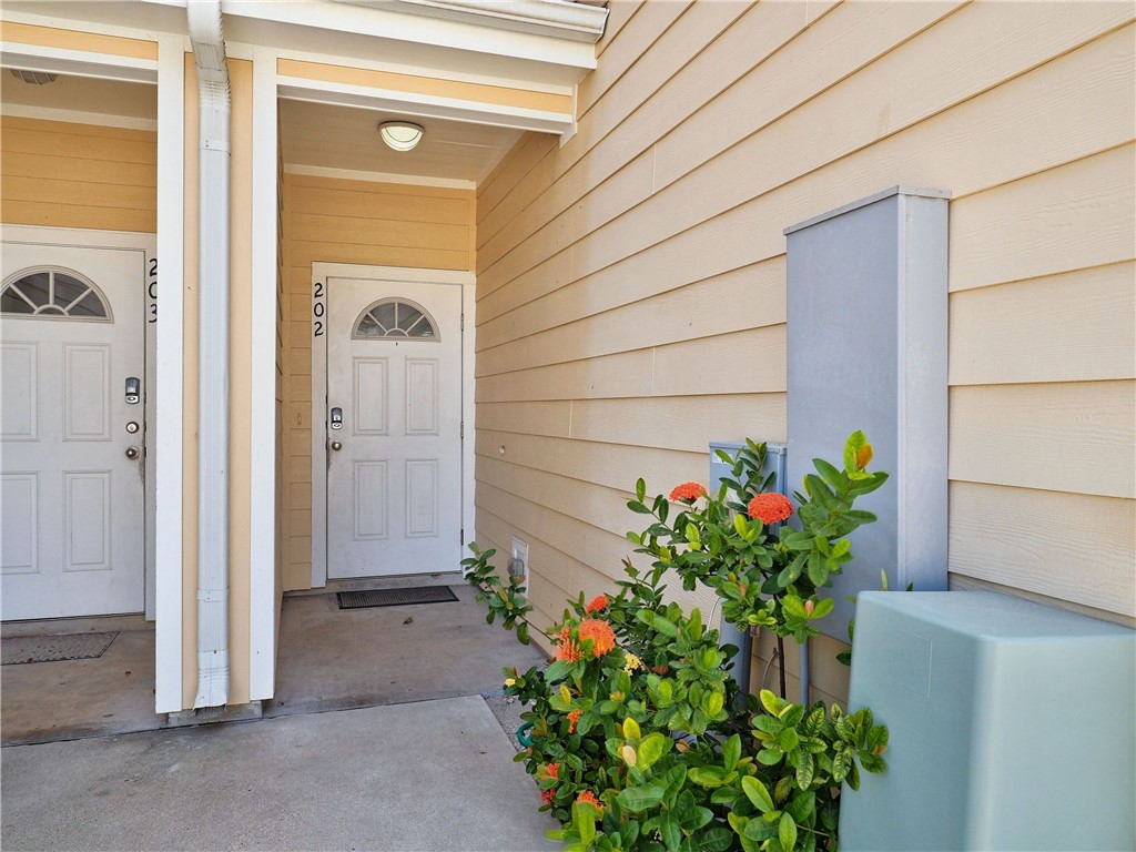 Unit 202 entrance with a white door, tan siding, and flowering shrubs along the walkway.