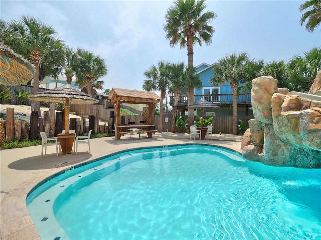 Outdoor swimming pool with a rock waterfall, palm trees, and shaded seating.