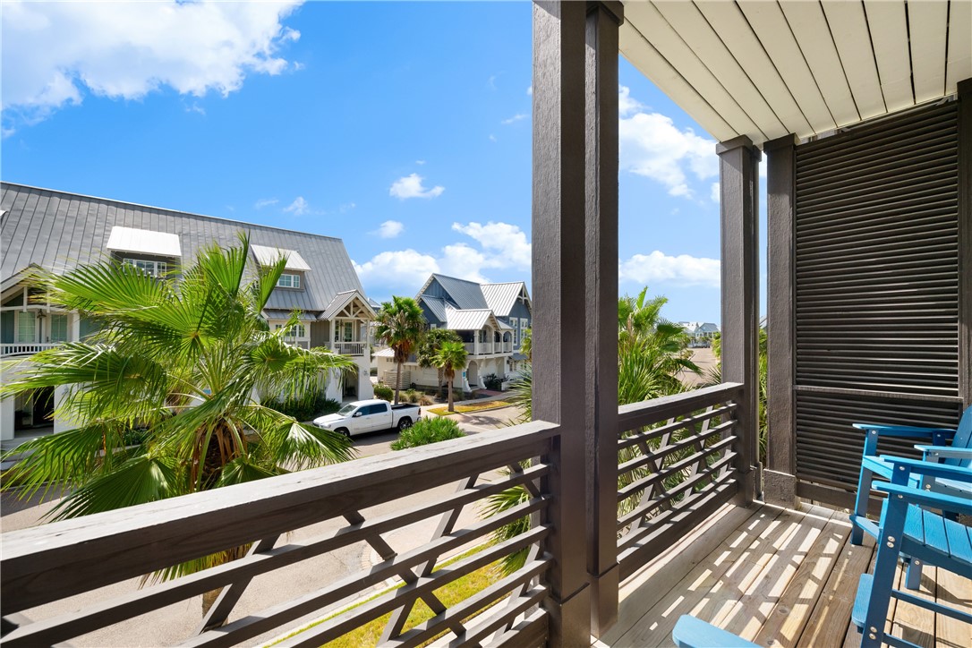 Covered wooden balcony with blue chairs overlooking palm trees and neighboring coastal-style homes.