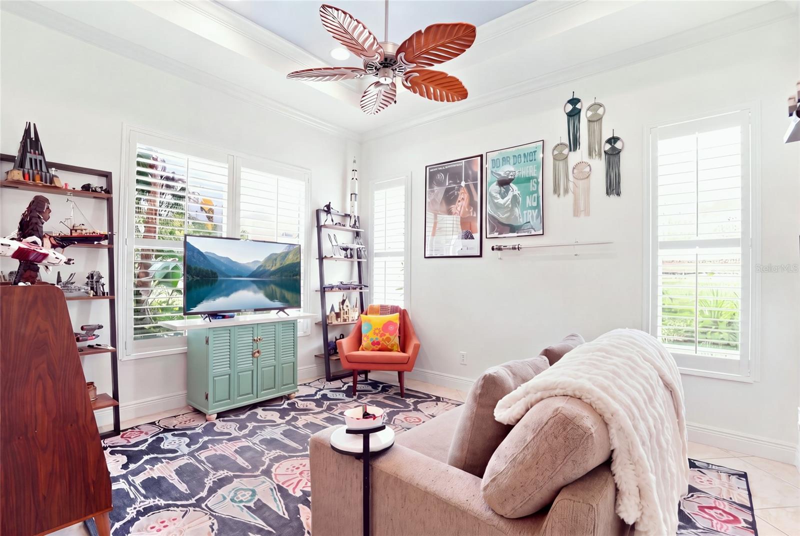 Bright living room with shuttered windows, colorful ceiling fan, television, and collectible display shelves.