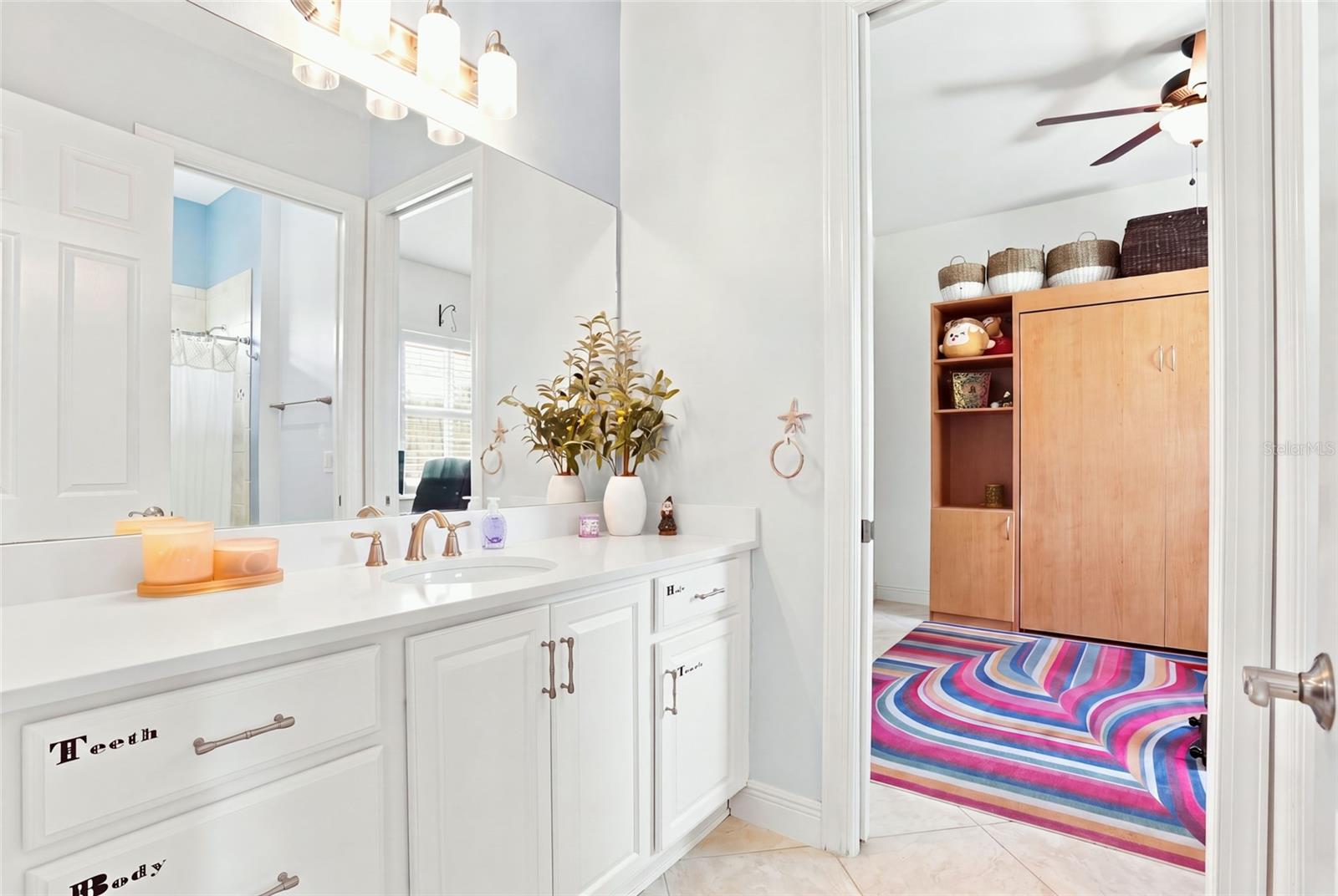 Bathroom vanity with white cabinetry and a large mirror, opening onto a room with a colorful rug.