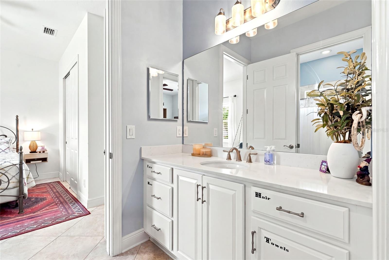 Bright bathroom vanity with large mirrors, labeled drawers, and a view into the adjoining bedroom.