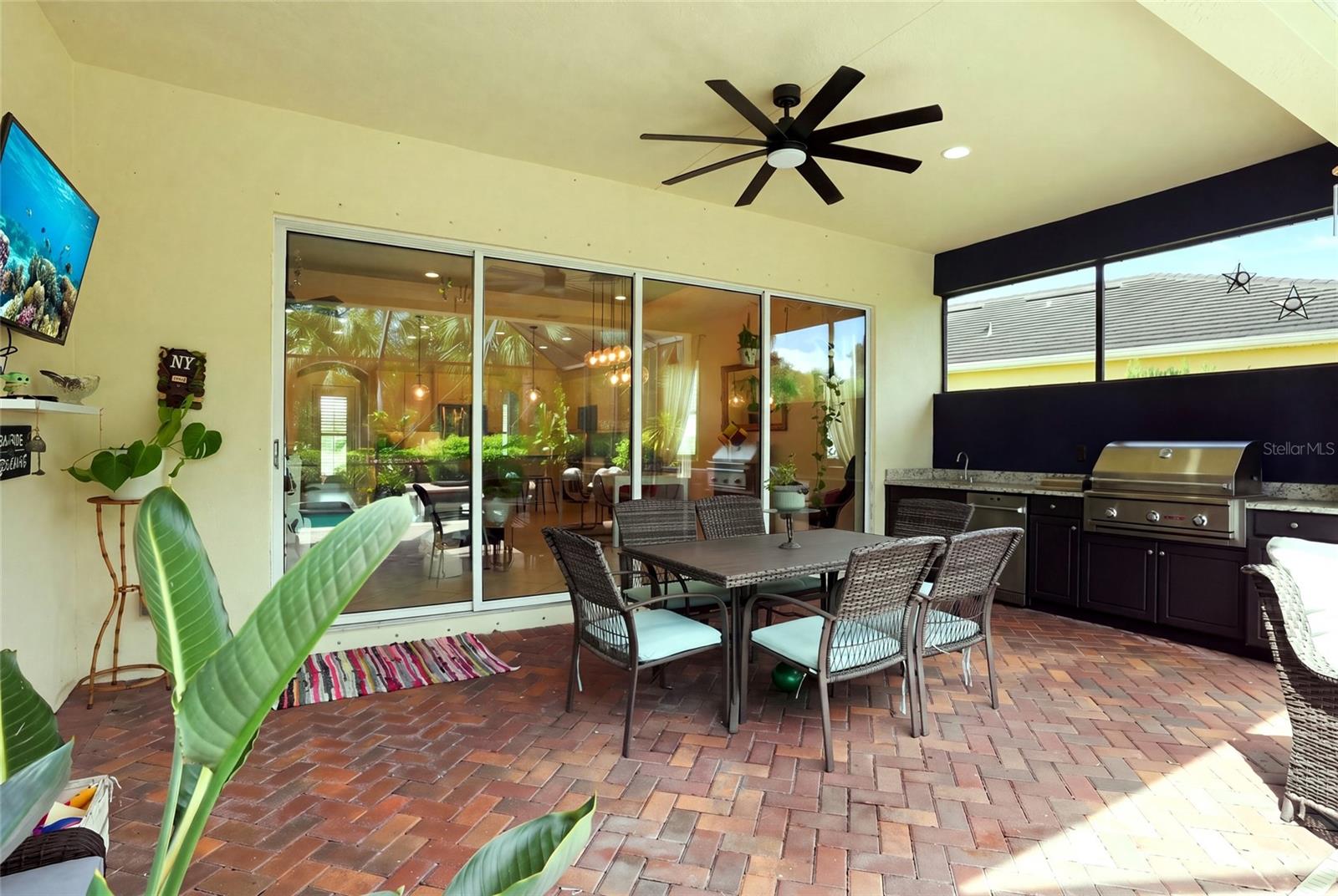 Covered patio with brick pavers, wicker dining set, built-in grill, and large sliding glass doors.