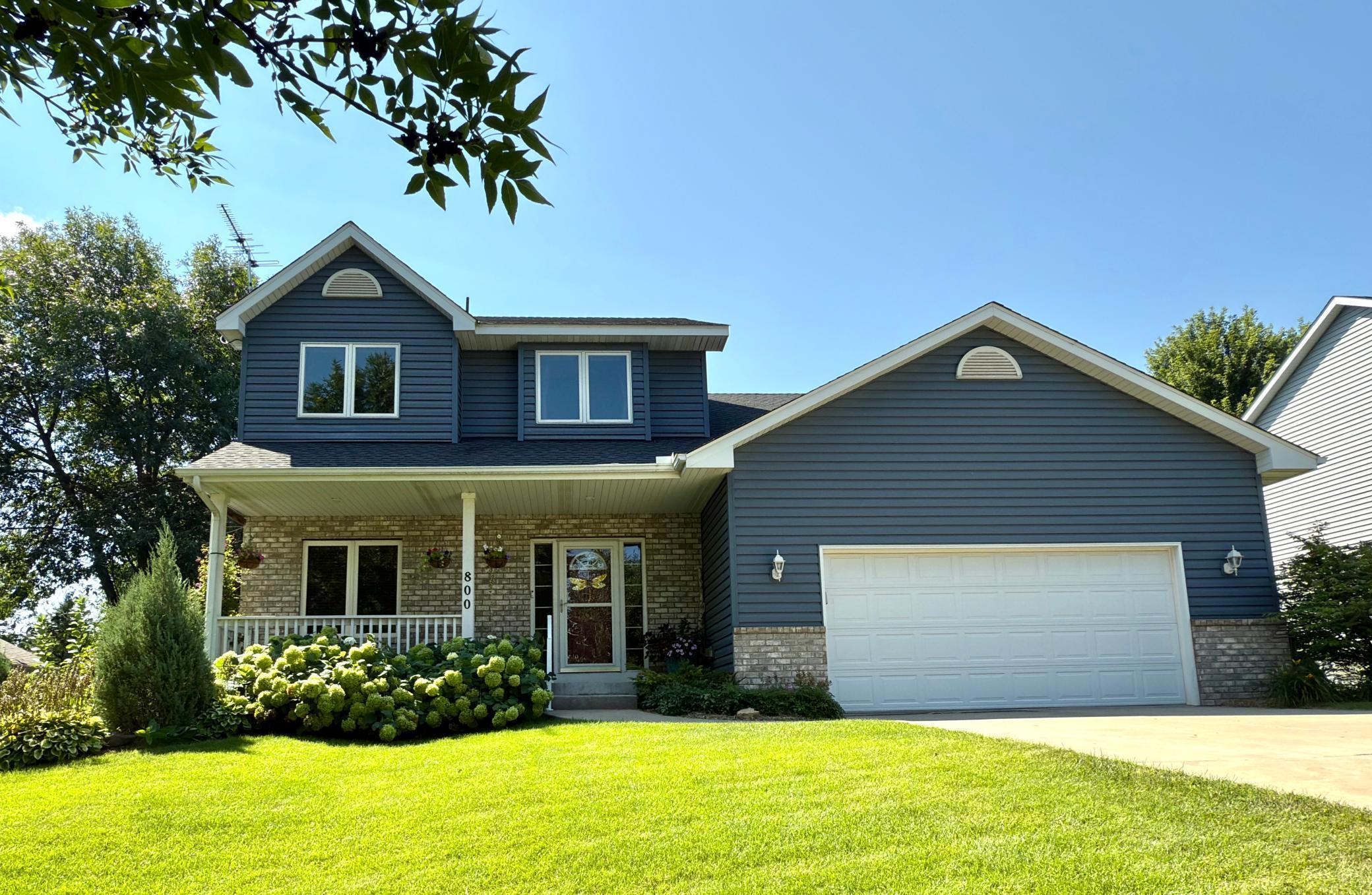Two-story blue-sided house with brick accents, a covered porch, attached garage, and landscaped front lawn.
